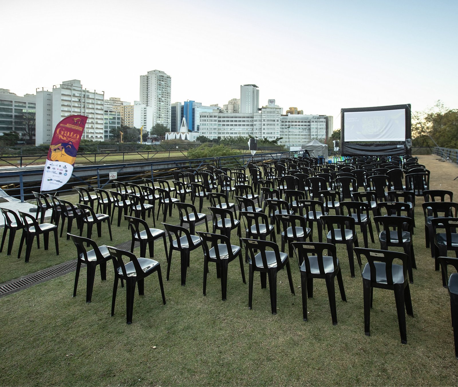 Fileiras de cadeiras vazias em uma sessão de cinema ao ar livre no jardim suspenso do Centro Cultural São Paulo, com o telão ao fundo e o horizonte da cidade