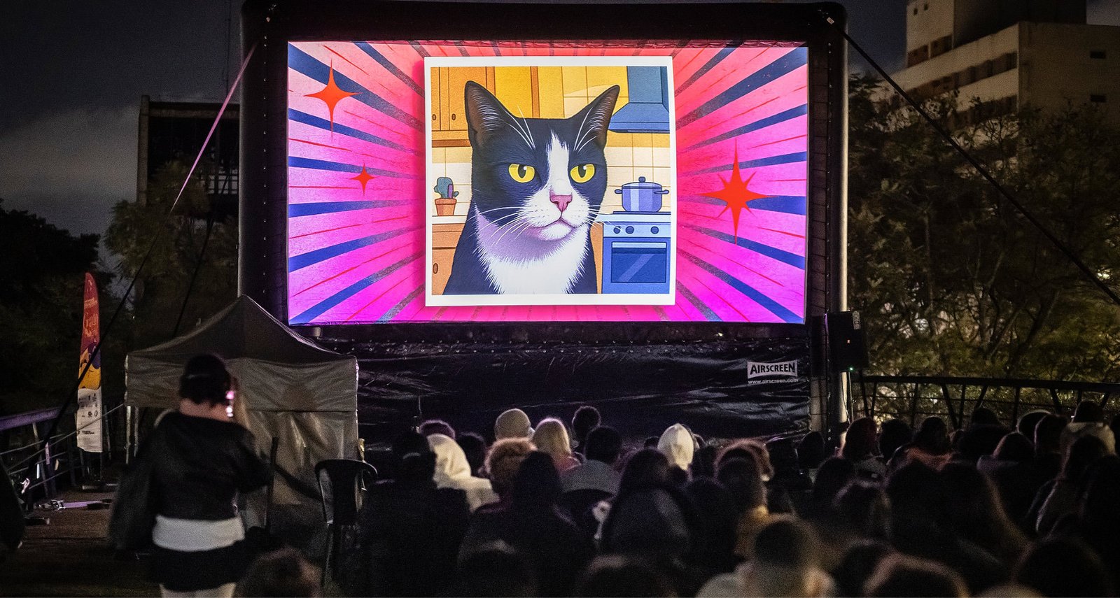 Público reunido para uma sessão de cinema ao ar livre à noite, assistindo a uma ilustração de gato no telão do CatVideoFest